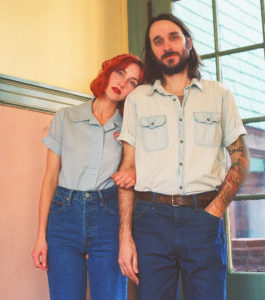 A young couple stand in front of a big window with a building seen through it. They are both white and wearing light-colored denim short-sleeved shirts and dark blue denim jeans. The woman stands to the left; she has chin-length slighly wavy red hair and leans her head on the man's shoulder. The man stands to the right and has dark brown shoulder-length hair and a beard/moustache.