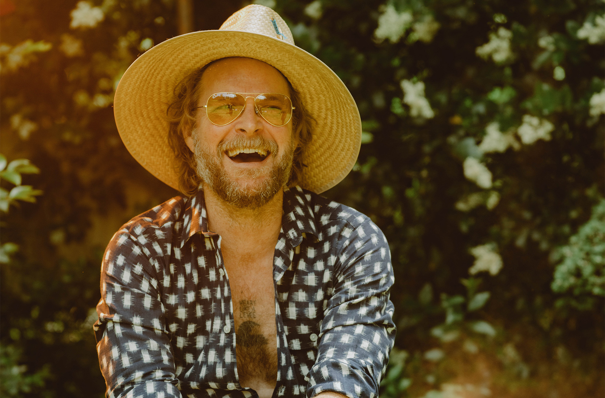 A photo of M.C. Taylor, band leader of the group Hiss Golden Messenger. He is outdoors in sunny weather wearing a hat and smiling.