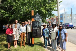 A photograph of 11 Black and white men and women standing around the DeFord Bailey TN Music Pathways marker. The marker is rectangular with info about and an image of DeFord Bailey; the TN Music Pathways guitar pick-shaped icon is above the main panel. 