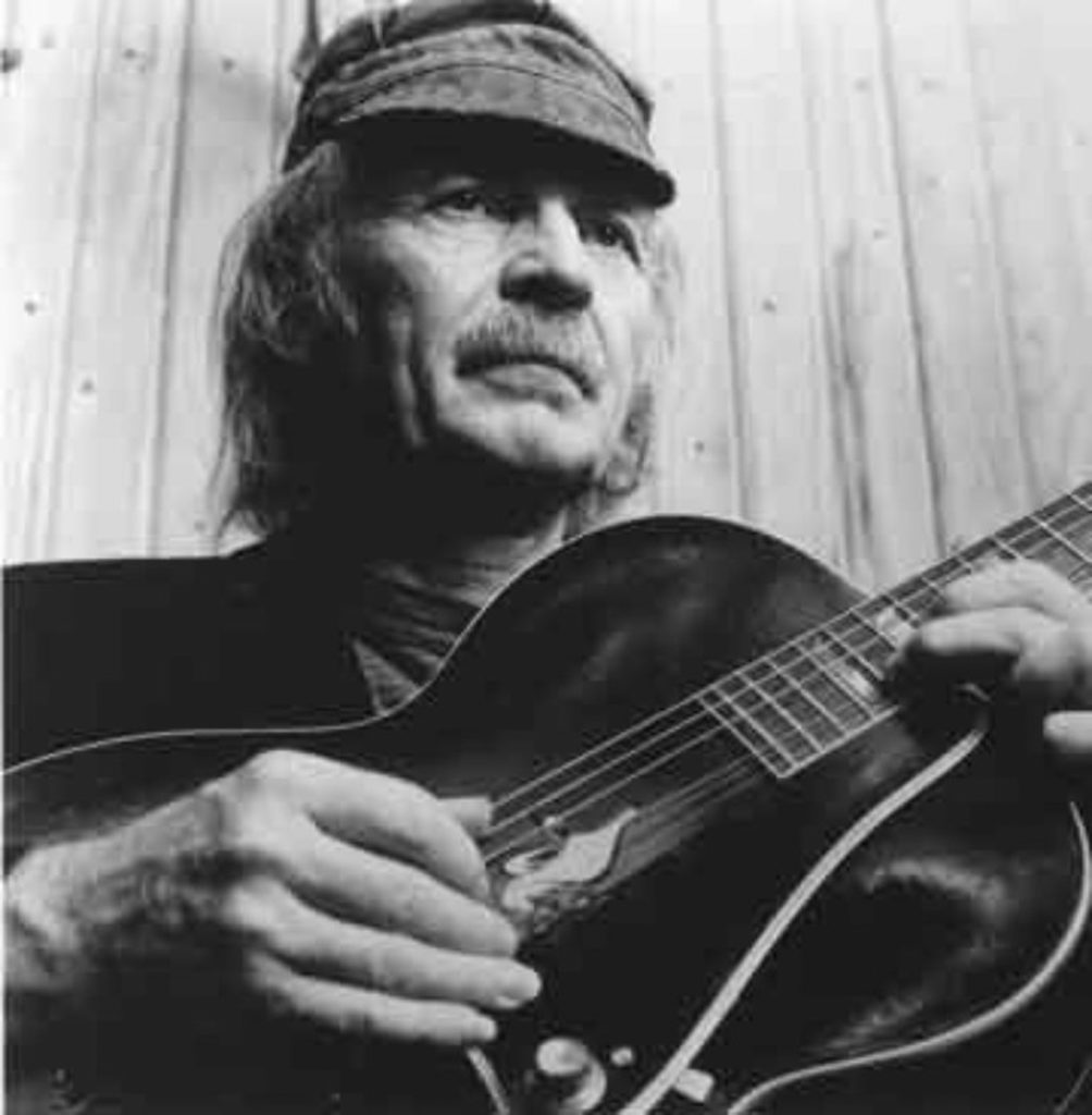 Black-and-white photograph of an older Michael Hurley wearing a cap and strumming a guitar.