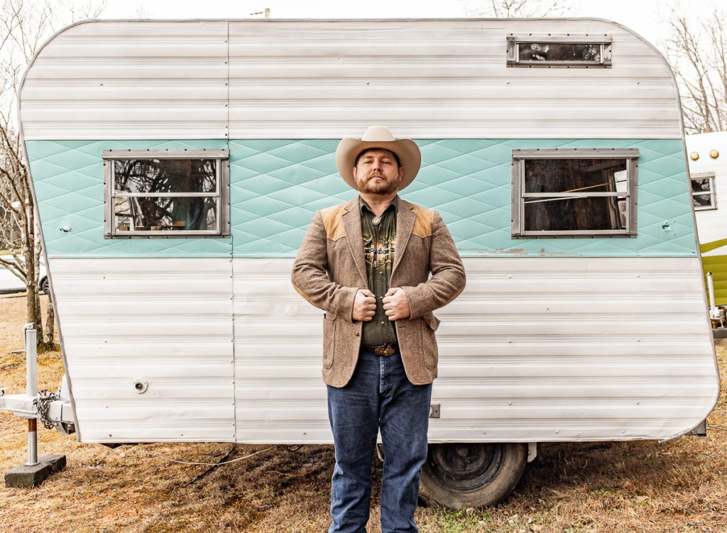 A white man with a brownish beard stands in front of an old pull-behind trailer. The trailer is white with an aqua stripe and two windows. The man is wearing a cowboy hat, a beige jacket with leather patches on the shoulder area, a patterned shirt and bolo tie, and jeans.