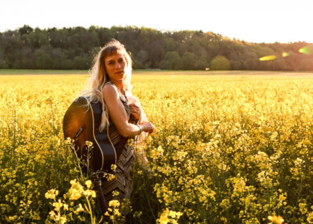 Anya Hinkle stands in a large field of yellow-flowering plants. A stand of green woods are seen in the background at the far end of the field. Anya is a white woman with long blonde hair and bangs. She carries a guitar on her back, and she is wearing a brown and black geometric-patterned dress.