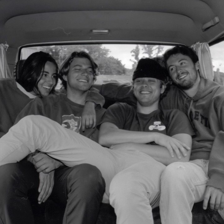  A black and white image of 4 young men in the back of a vehicle. The men are all smiling and have their arms around one another. The men are wearing t-shirts and hoodies. 