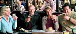 Cast members of the film Shaun of the Dead sitting in a booth at The Winchester pub raising up pints of beer and smiling.