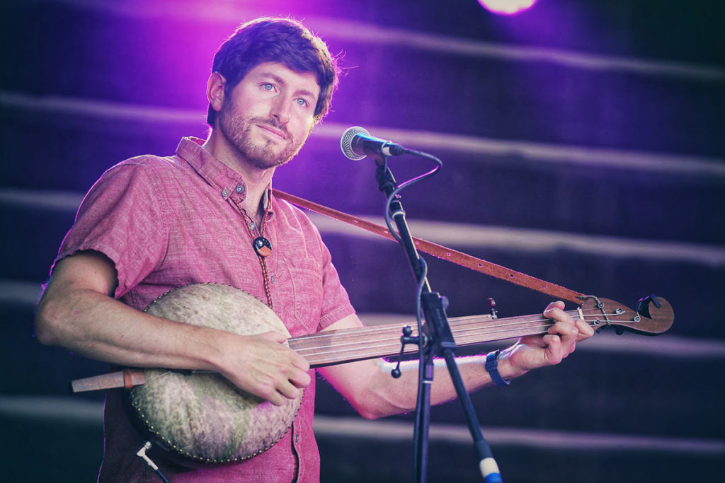 Image shows a white man standing on stage. He has brown hair, blue eyes, and a short brown beard. He is wearing a pink short-sleeved shirt and holds a big round gourd banjo. The stage lights behind him are a purple color.