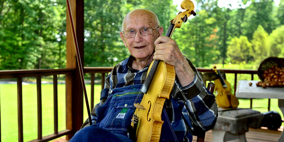 The image is of an elderly white man sitting on a wooden porch with trees. He is wearing glasses, a blue plaid shirt, jean overalls, and he is holding a yellow-wood fiddle and bow.