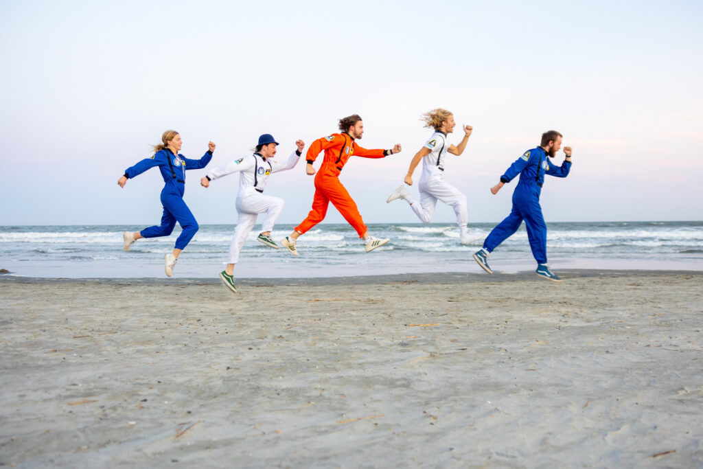 Five people are wearing blue, or white, or orange jumpsuits and are jumping in mid air on a beach. 