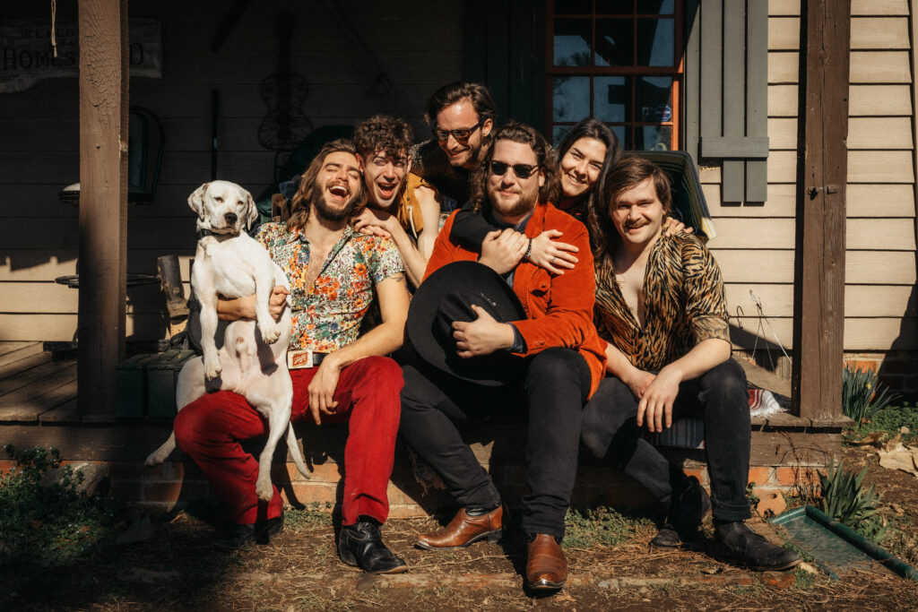 A fun group image of the band Holler Roller. The group consists of six men and one woman, they are all sitting on a porch with their arms around each other. The man on the far left is holding a white dog in his lap. Everyone is wearing colorful clothing and smiling and laughing. 