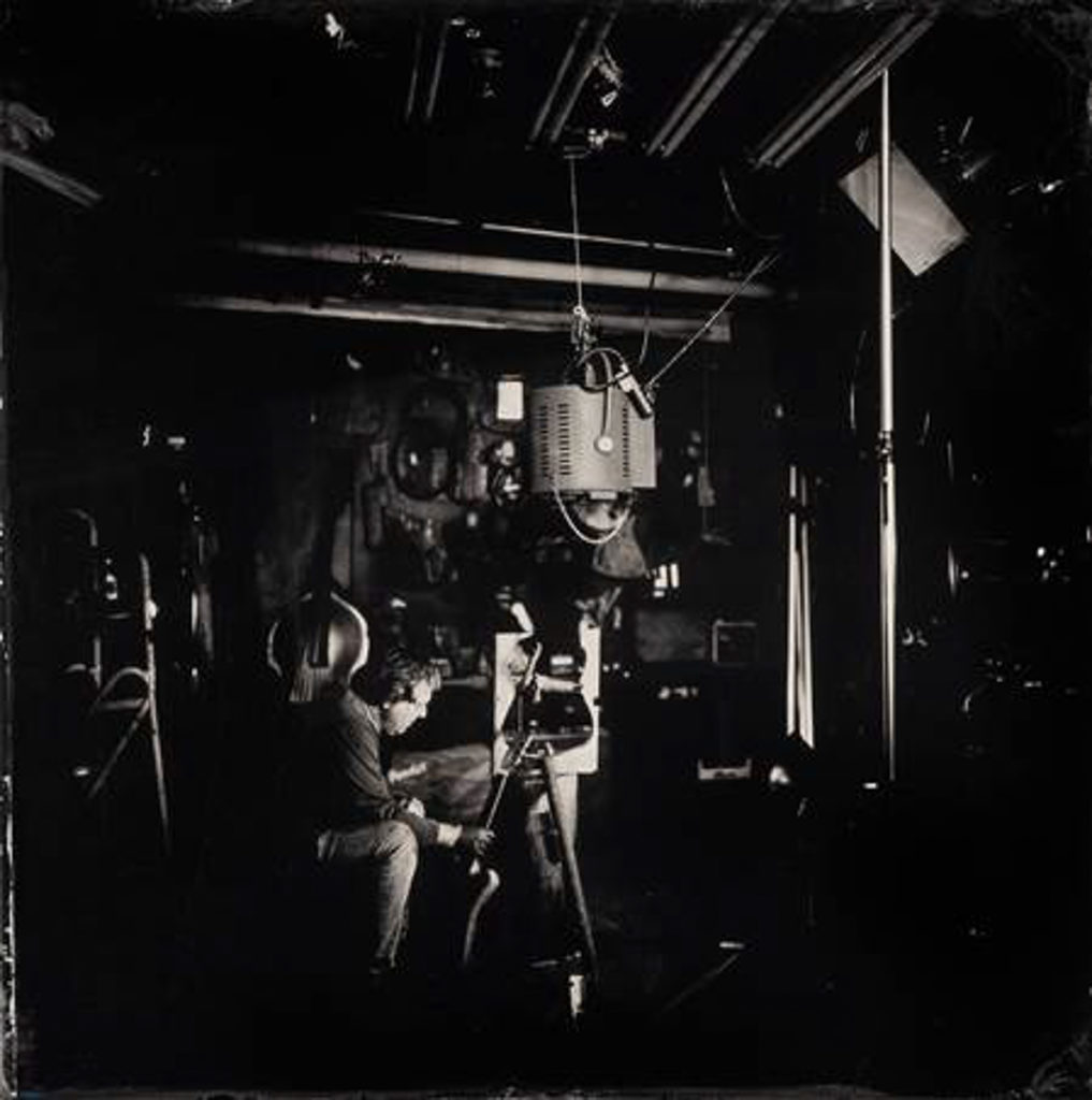 A dramatically lit black-and-white photo showing a white man sitting in front of some large camera equipment. In the background, you can see several stringed instruments leaning against and hanging on the wall.