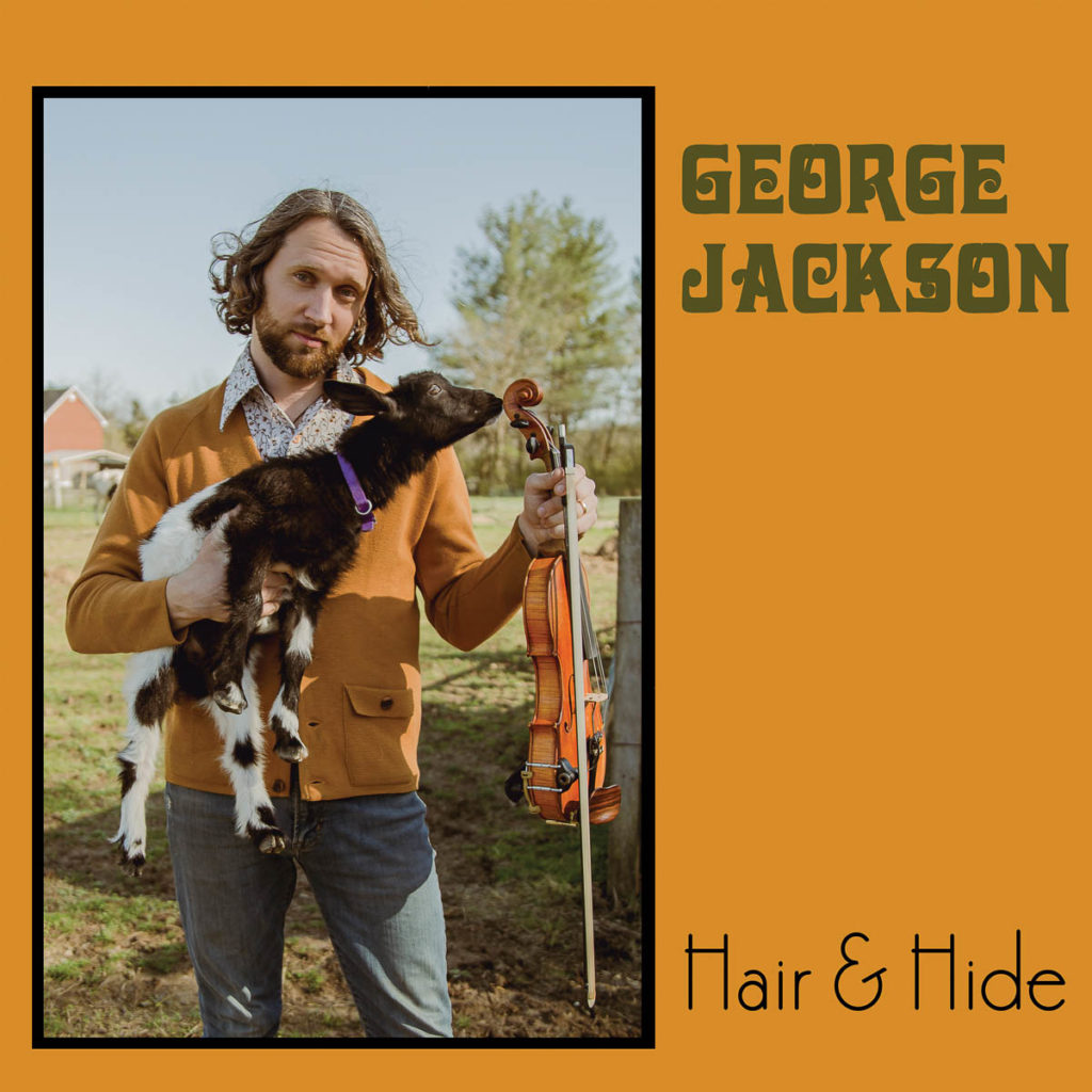 The cover image shows George Jackson standing in a pasture with a barn or farmhouse behind him. He is a white man with slightly longer wavy hair and a beard. He is wearing a white patterned button-down shirt with a brown or ochre-colored cardigan and jeans. He holds a small black and white goat in one hand and a fiddle in the other. 