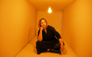 A photo of Jade Bird sitting in a box. There is a single light bulb hanging from the center above her head.