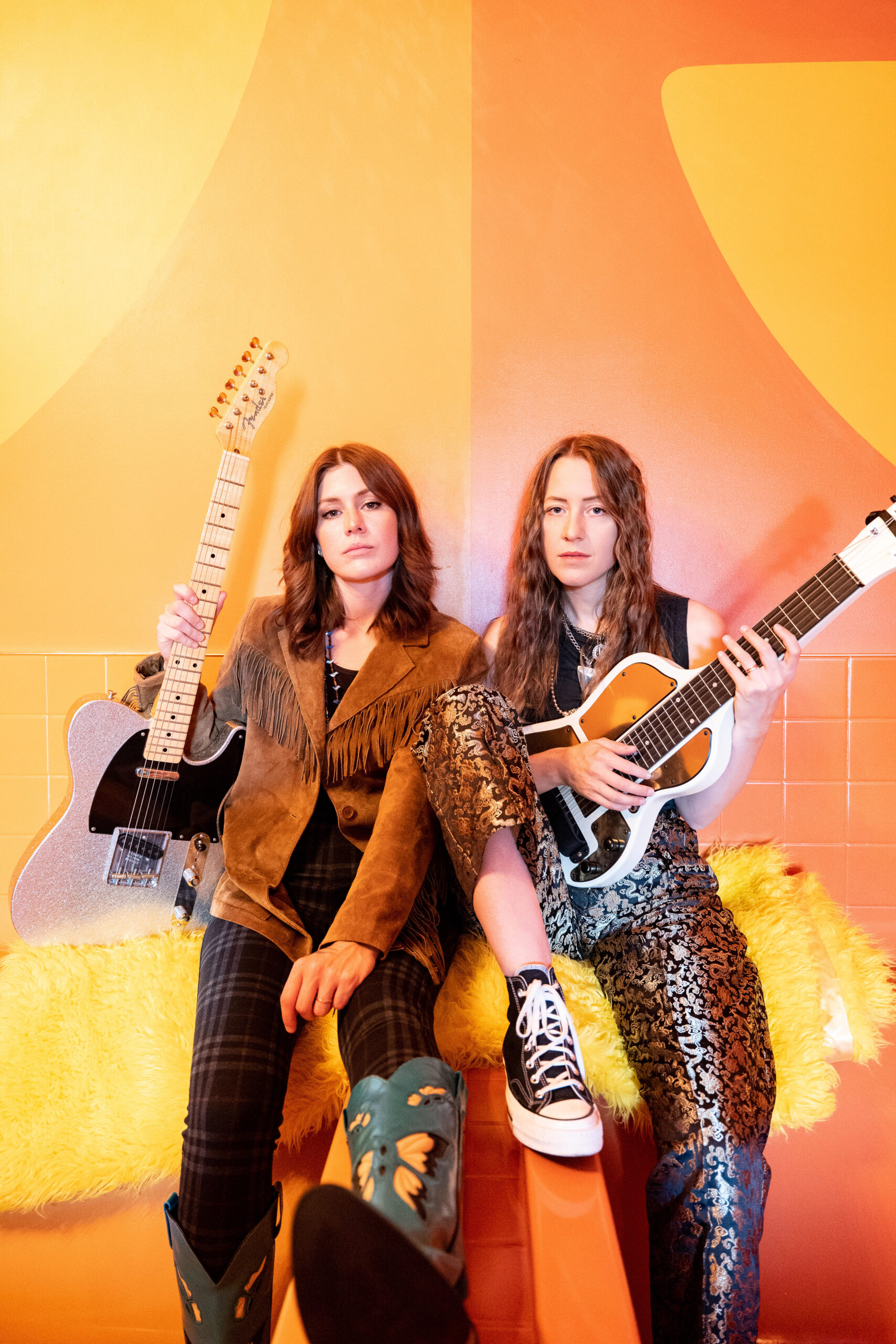 Promotional image of the duo Rebecca and Megan Lovell of Larkin Poe. Two women are facing the camera and holding instruments. The women on the left is holding a light pink guitar with glitter and wearing a brown fringe western jacket. The woman on the right is holding an instrument and wearing a black sleeveless shirt with shiny patterned pants. Both women are sitting on a fuzzy yellow blanket in front of a colorful orange and yellow background. 