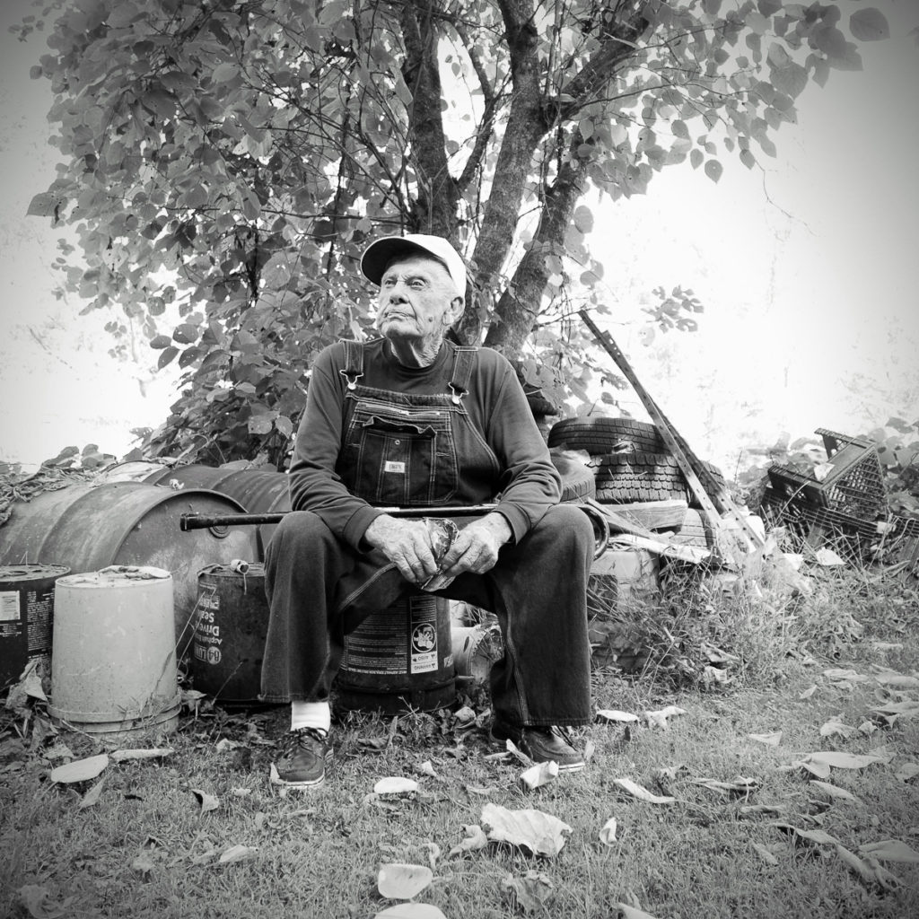 An old white man wearing a dark long-sleeved shirt with jean overalls and a white baseball / trucker-style cap. He is clean-shaven. He sits in front of some old oil barrels, tires, and other items in a yard area.