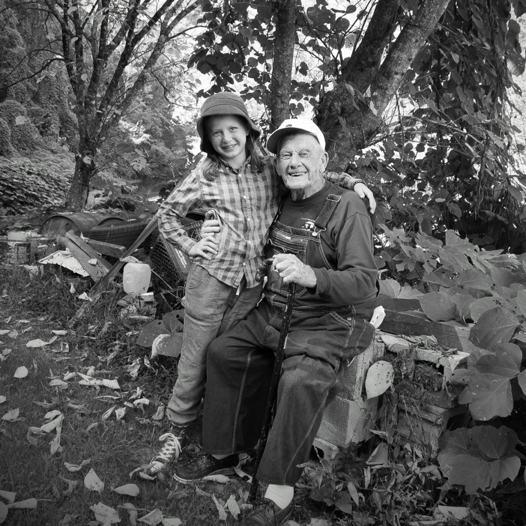 A young white girl with blondish hair and wearing a plaid long-sleeve shirt, light-colored pants, a bucket hat, and converse sneakers has her arm around an old white man in a dark long-sleeved shirt and overalls. He is seated and wearing a white baseball cap and holding a wooden cane. She is standing beside him. Trees and an old wall can be seen in the picture too.