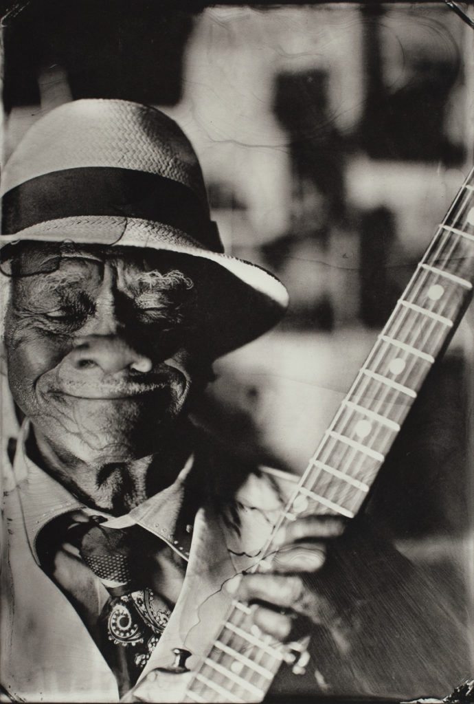 A Black man is wearing a light colored fedora-style hat with a black sash on it; he also wears a light-colored suit and a tie decorated with paisleys. He has one hand on the neck of a banjo. His eyes are closed and he has a beautific smile on his face.
