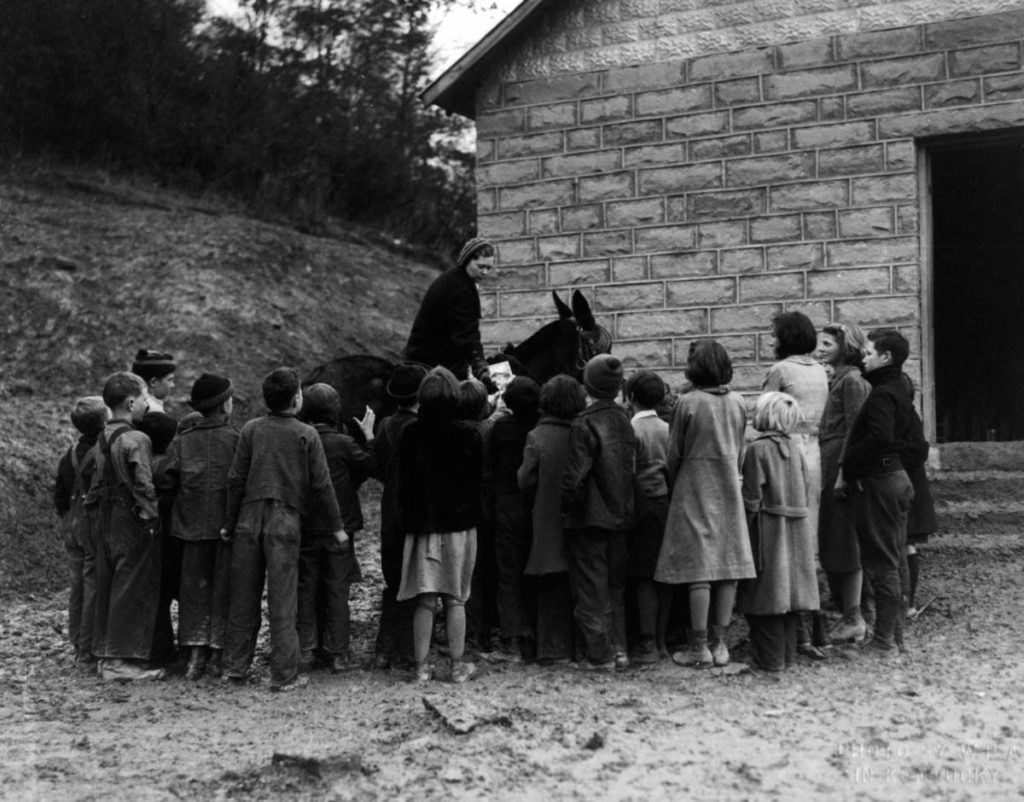 A WPA archive image showing a female librarian on a pack horse or mule surrounded by a cluster of children, waiting for her to give out books. They are in front of a small stone schoolhouse.