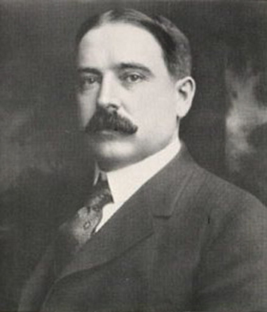 A black-and-white portrait of a white man with dark hair and a big moustache. He is wearing a dark suit and patterned tie.