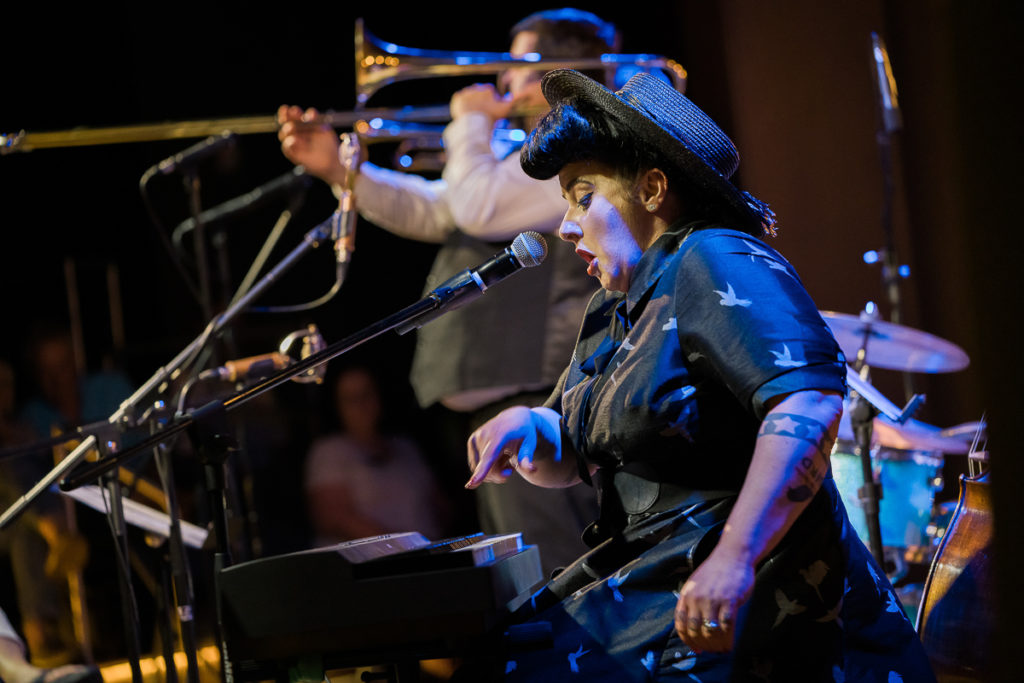 A close-up of Davina playing the keyboards, dressed all in black and with a hat. The band's trombonist is seen in the background.